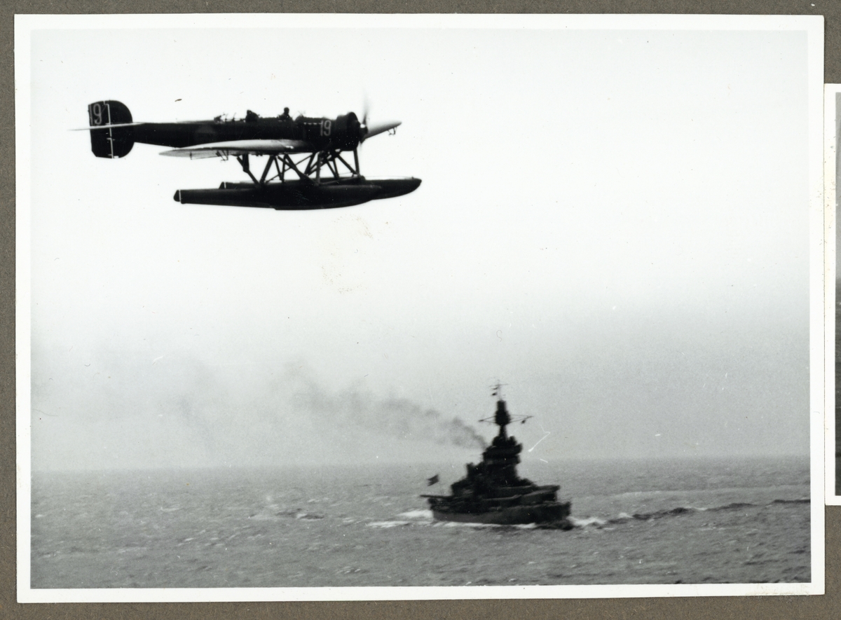 Bilden visar en Heinkel HE 5 sjöflygplan som flyger över havet. Under den syns ett örlogsfartyg.