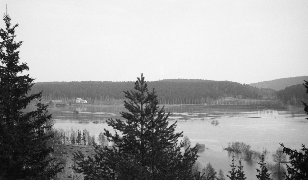 Flom i Glomma ved Rena i Åmot i Hedmark 6. mai 1934. Fotografiet er tatt fra lia på østsida av vassdraget, like nordvest for Åmot prestegard med kameraet vendt østover mot den oversvømte dalbotnen. På motsatt side av elva ser vi over mot gardstunet på jord- og skogeiendommen Kilde.