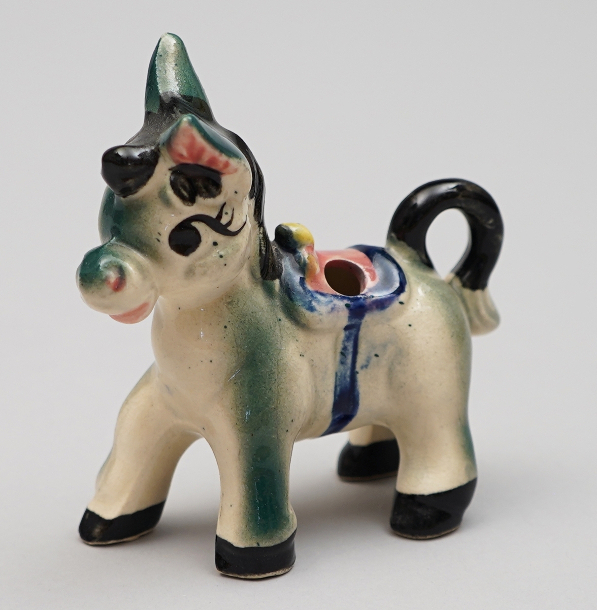 Eselfigur i steingods, måla dekor. Eit hol på kvar side av sadelen.
