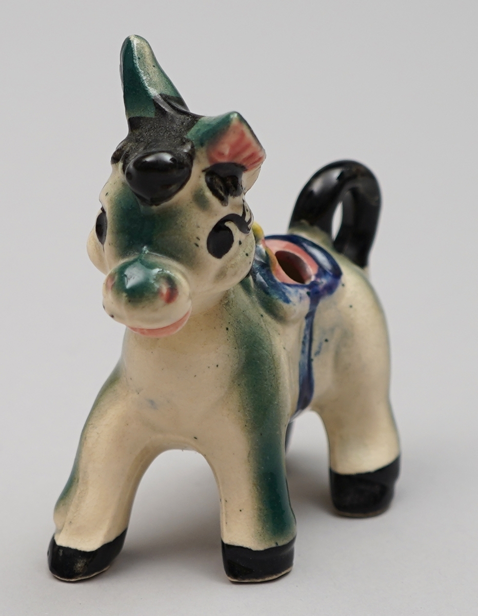 Eselfigur i steingods, måla dekor. Eit hol på kvar side av sadelen.