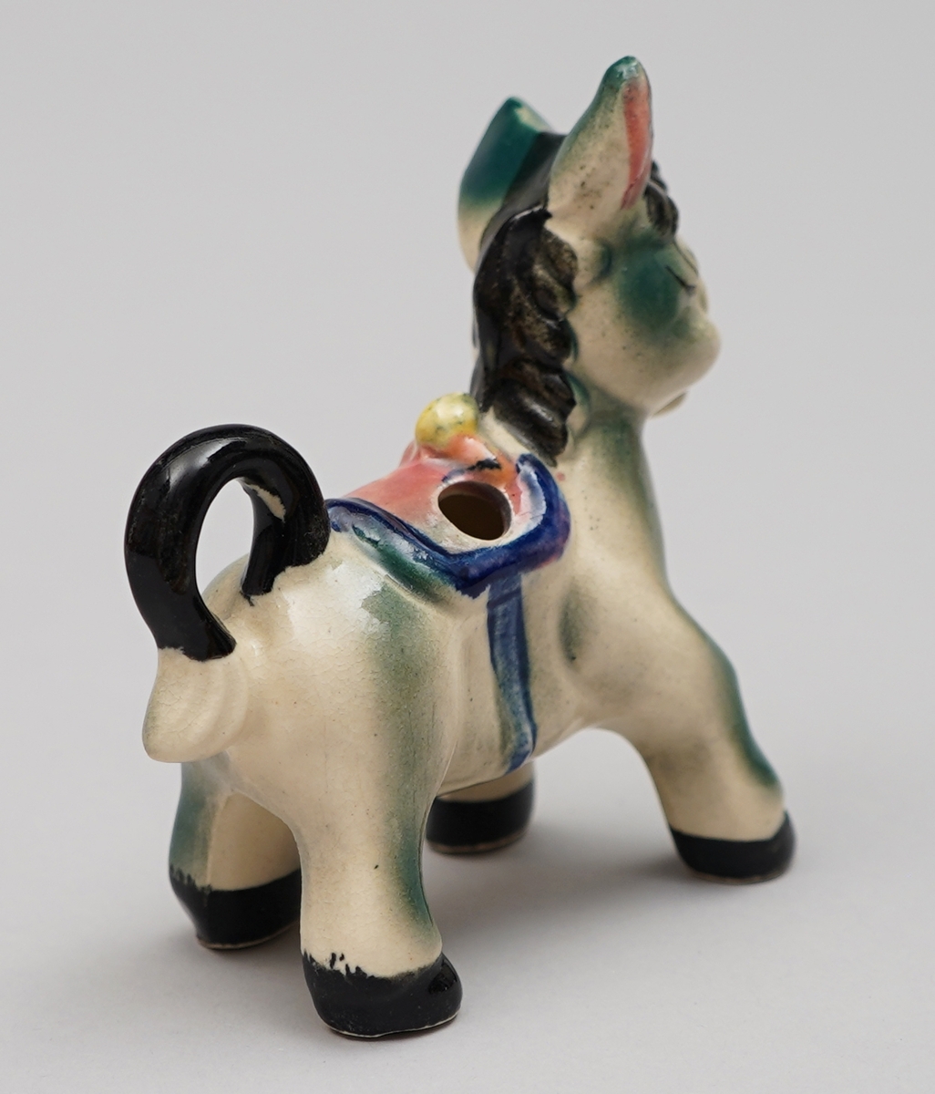 Eselfigur i steingods, måla dekor. Eit hol på kvar side av sadelen.