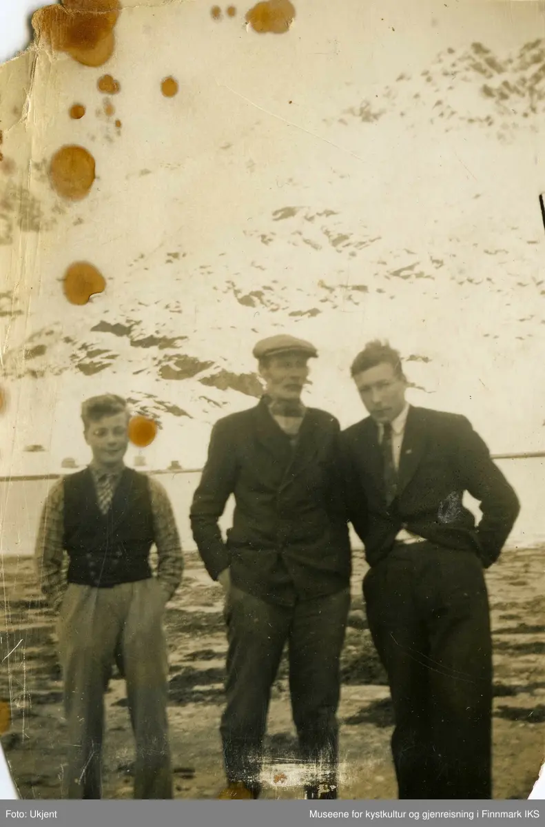Gruppebilde av tre men. De står foran havet og snedekket fjell. Alle har på seg finklær og de to til høyre har på seg dressjakker. Mannen i midten har sixpence på seg. Alle tre har hendene i lommene. Opptakssted og personene, samt fotograf er ukjent.