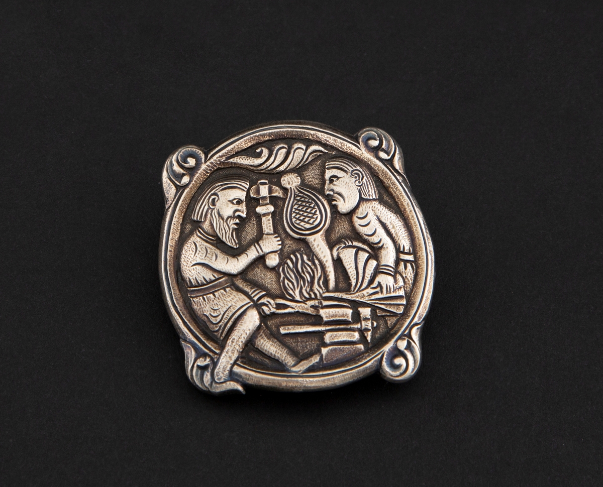 Sirkulær spenne i støpt sølv med siselert detaljering. Fremstilling av Regin som smir sverdet i lavt relieff. Motivet er hentet fra Hyllestadportalen, og viser scener fra sagaen om Sigurd Favnesbane. Spennens hjørner er markert med fire krumme løv. På baksiden er det en krok.