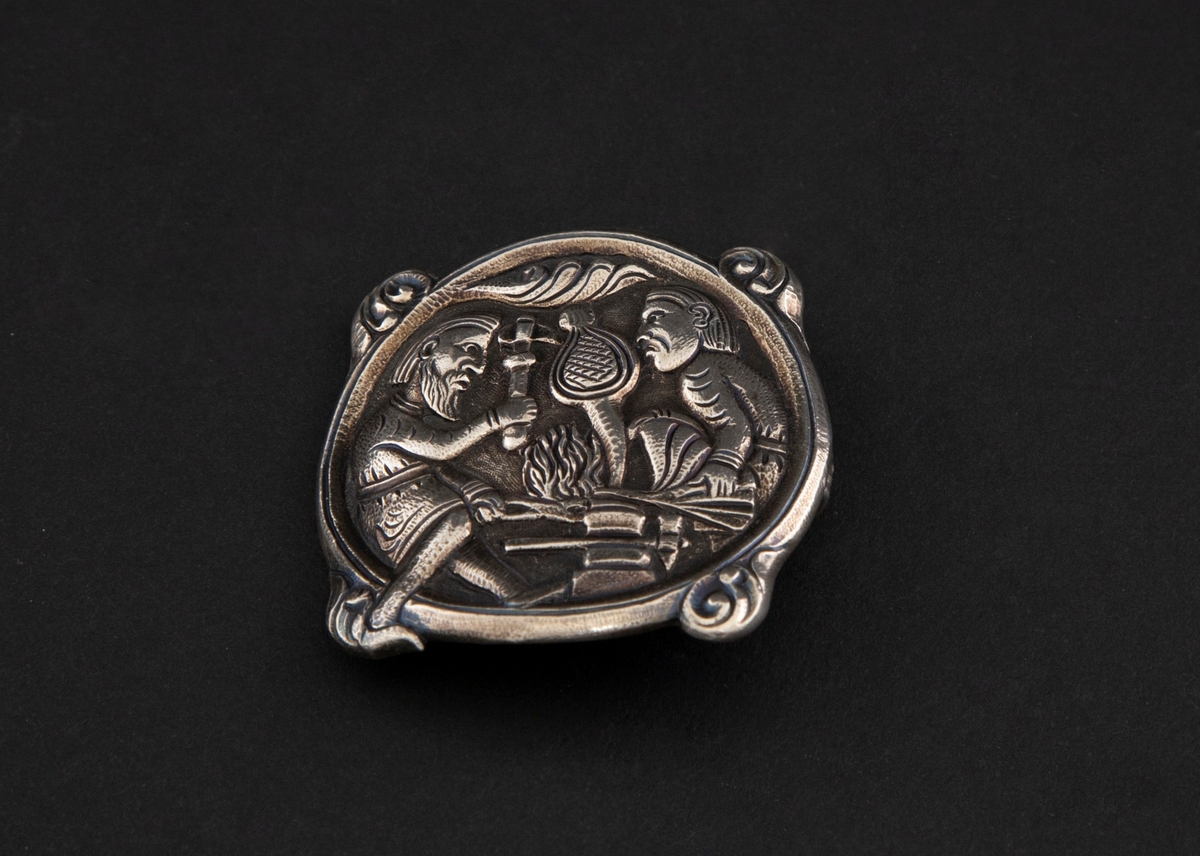 Sirkulær spenne i støpt sølv med siselert detaljering. Fremstilling av Regin som smir sverdet i lavt relieff. Motivet er hentet fra Hyllestadportalen, og viser scener fra sagaen om Sigurd Favnesbane. Spennens hjørner er markert med fire krumme løv. På baksiden er det en krok.