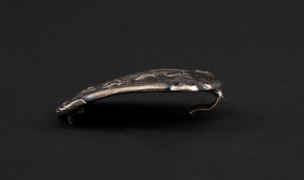 Sirkulær spenne i støpt sølv med siselert detaljering. Fremstilling av Regin som smir sverdet i lavt relieff. Motivet er hentet fra Hyllestadportalen, og viser scener fra sagaen om Sigurd Favnesbane. Spennens hjørner er markert med fire krumme løv. På baksiden er det en krok.