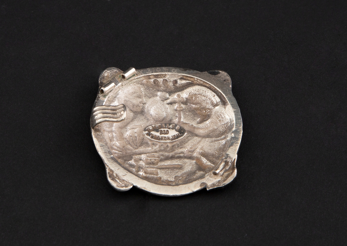 Sirkulær spenne i støpt sølv med siselert detaljering. Fremstilling av Regin som smir sverdet i lavt relieff. Motivet er hentet fra Hyllestadportalen, og viser scener fra sagaen om Sigurd Favnesbane. Spennens hjørner er markert med fire krumme løv. På baksiden er det en krok.