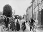 Tre flickor och två pojkar står bredvid sina cyklar på Brahegatan strax norr om torget i riktning norrut. De står utanför en byggnad med texten 
