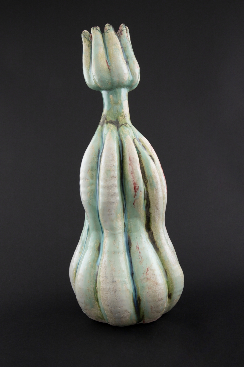 Håndmodellert skulptur i glasert steingods med oppreist holdning. Den har en organisk og asymmetrisk kalebassform som ender i en tulipanform med halvåpne kronblader. Keramikkskulpturen inngår i en installasjon bestående av 26 deler i ulike størrelser og med individuell utforming. De asymmetriske variasjonene gir et inntrykk av bevegelse, og den lett sykelige fargepaletten gir et uhyggelig og grotesk preg. Samtlige deler minner om kjøttetende planter.