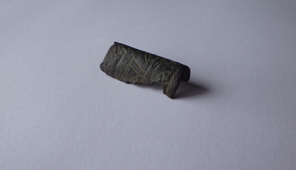 MALV: L. 25 MM, BÅNDFORMET FRAGMENT SOM ER BØYET. OVERSIDEN HARINNPRESSET DEKOR. DEN ENE ENDEN BEARBEIDET. 