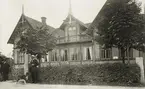 Bostadshus, Åseda, ca 1912. En man står vid grinden med en hund. På husväggen syns reklamskyltar 
för försäkringsbolaget Skandia.