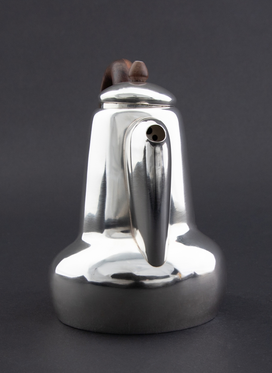 Kanne i sølv med håndtak av palisander. Ovalformet korpus samt høyreist hals med fremtrukket tut. Mykt buet håndtak samt lokknapp i palisander. Samtlige deler preges av mykt avrundede kanter. Innsiden er forgylt.