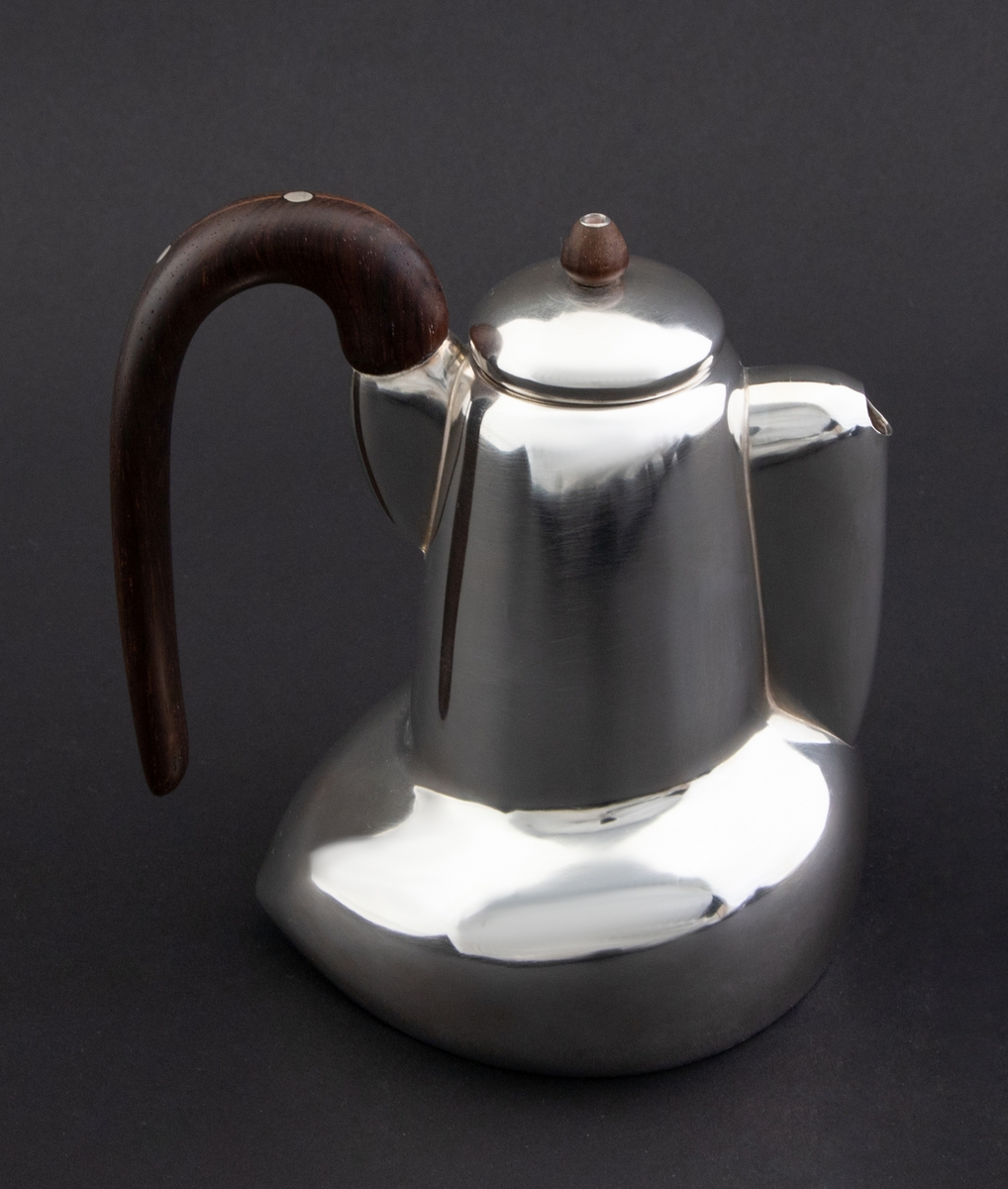 Kanne i sølv med håndtak av palisander. Ovalformet korpus samt høyreist hals med fremtrukket tut. Mykt buet håndtak samt lokknapp i palisander. Samtlige deler preges av mykt avrundede kanter. Innsiden er forgylt.