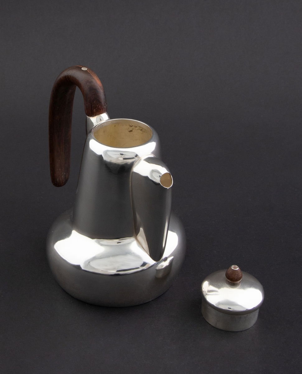 Kanne i sølv med håndtak av palisander. Ovalformet korpus samt høyreist hals med fremtrukket tut. Mykt buet håndtak samt lokknapp i palisander. Samtlige deler preges av mykt avrundede kanter. Innsiden er forgylt.