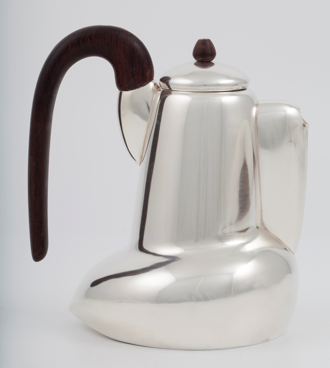 Kanne i sølv med håndtak av palisander. Ovalformet korpus samt høyreist hals med fremtrukket tut. Mykt buet håndtak samt lokknapp i palisander. Samtlige deler preges av mykt avrundede kanter. Innsiden er forgylt.