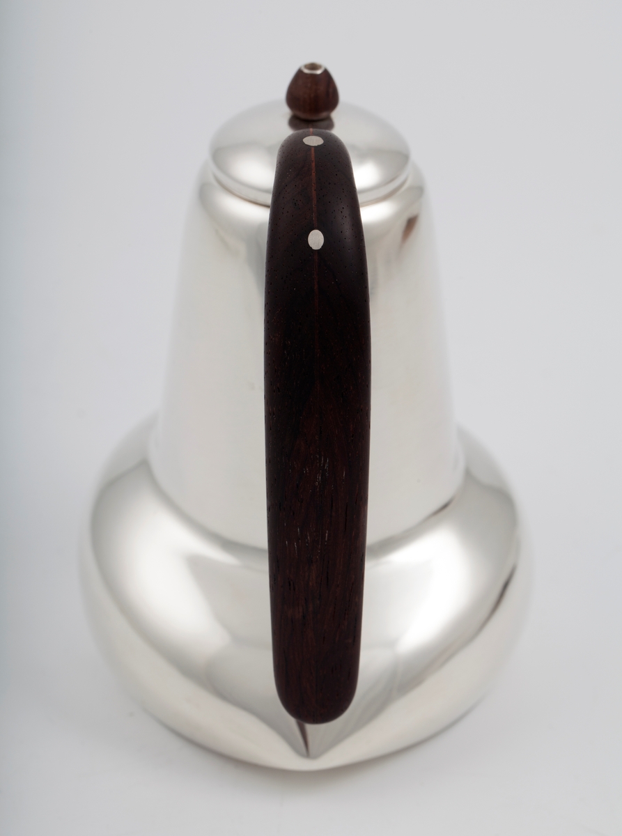 Kanne i sølv med håndtak av palisander. Ovalformet korpus samt høyreist hals med fremtrukket tut. Mykt buet håndtak samt lokknapp i palisander. Samtlige deler preges av mykt avrundede kanter. Innsiden er forgylt.