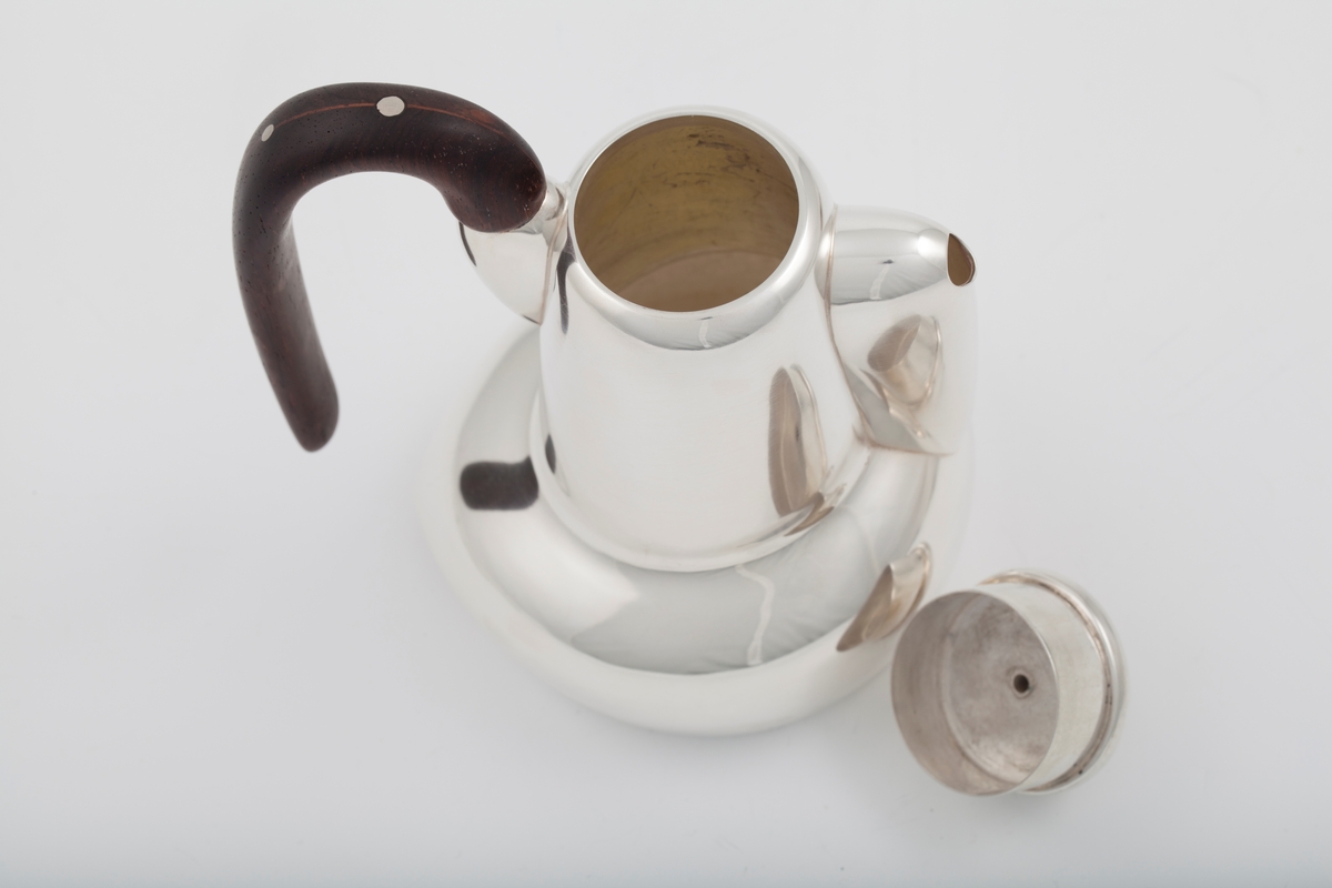 Kanne i sølv med håndtak av palisander. Ovalformet korpus samt høyreist hals med fremtrukket tut. Mykt buet håndtak samt lokknapp i palisander. Samtlige deler preges av mykt avrundede kanter. Innsiden er forgylt.