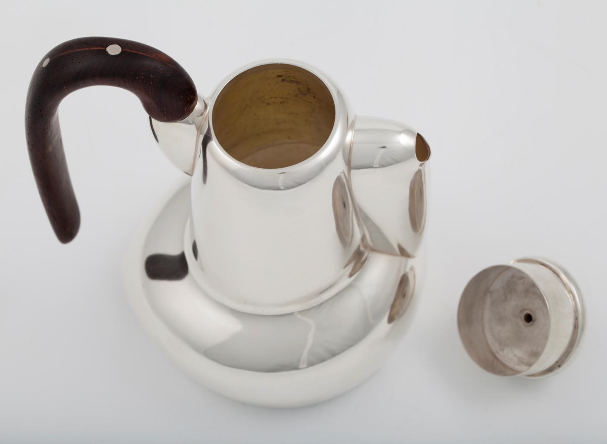 Kanne i sølv med håndtak av palisander. Ovalformet korpus samt høyreist hals med fremtrukket tut. Mykt buet håndtak samt lokknapp i palisander. Samtlige deler preges av mykt avrundede kanter. Innsiden er forgylt.