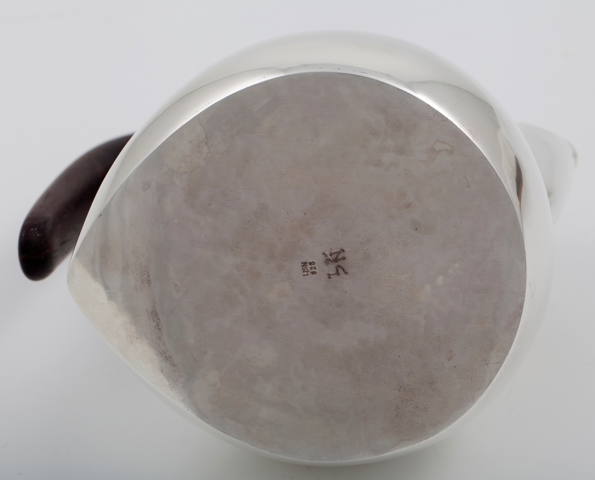 Kanne i sølv med håndtak av palisander. Ovalformet korpus samt høyreist hals med fremtrukket tut. Mykt buet håndtak samt lokknapp i palisander. Samtlige deler preges av mykt avrundede kanter. Innsiden er forgylt.