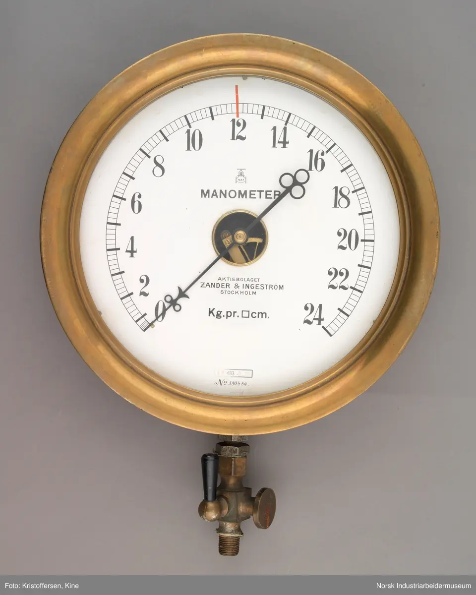 Manometer. Sirkulært måleinsturment i messingskall. Buet innhakk i messingen rundt glasset. Måleskiven med pil viser tall fra 0-24. Pilen har to sirkler i tupp og tre sirkler i enden. Åpent inn til tannhjulene i midten hvor pilen er festet.