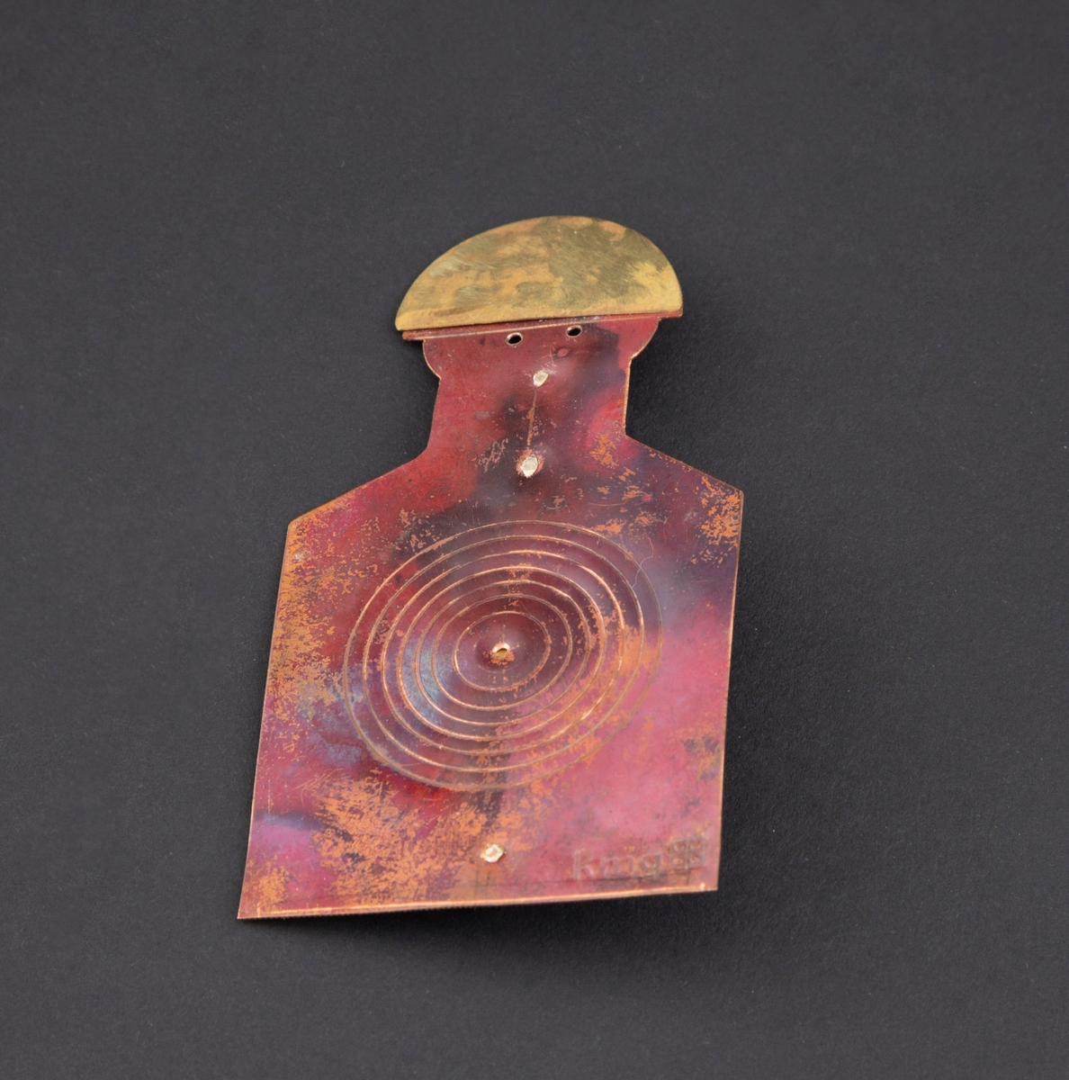 Brosje som forestiller en person med blink på brystet. Personen har gullhat og rødlig kobberkrop. Blink på brystet.