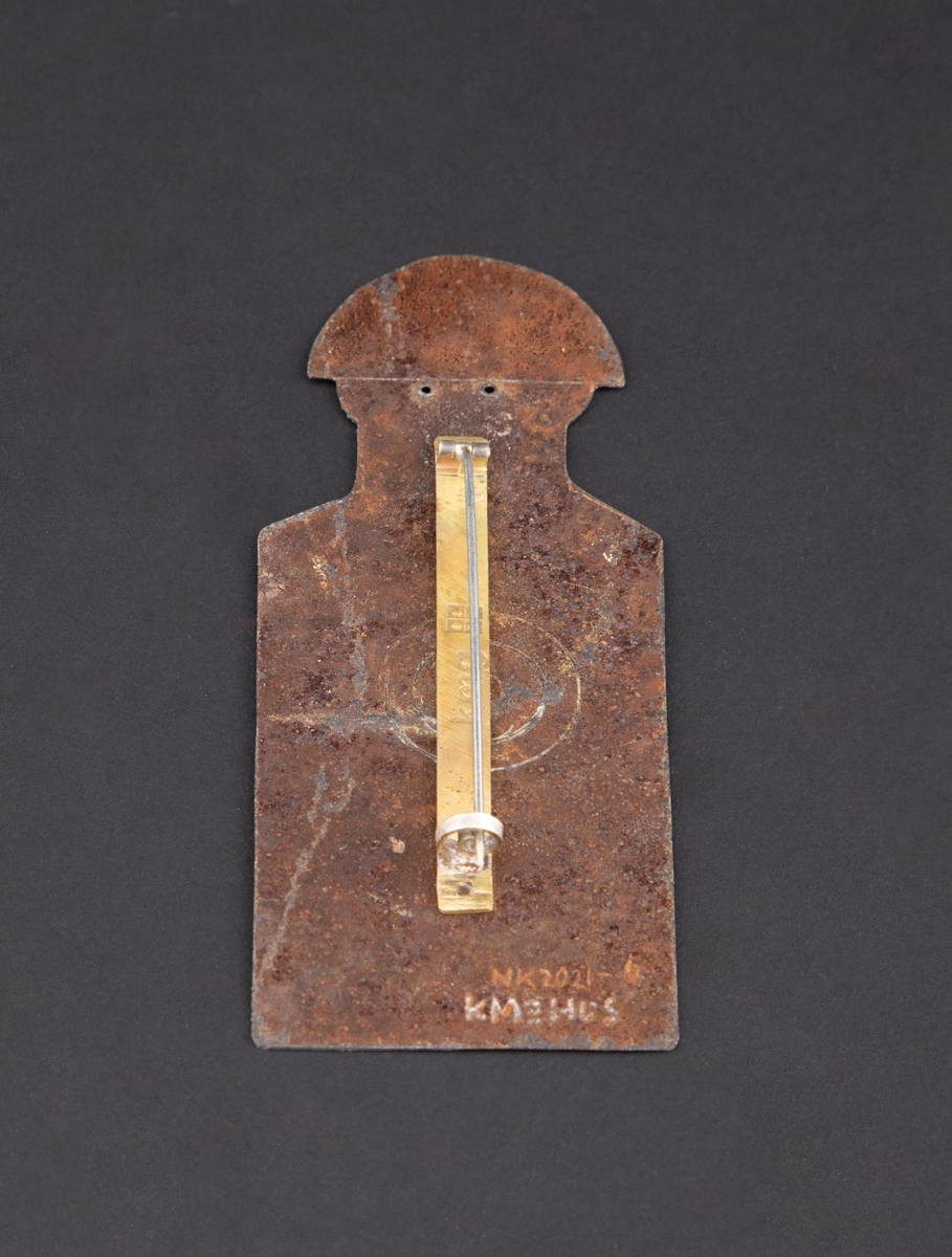 Brosje som forestiller en person med blink på brystet. Personen har rød hat og rustet kropp. Rød blink på brystet.