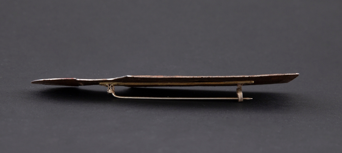 Brosje som forestiller en person med blink på brystet. Personen har rød hat og rustet kropp. Rød blink på brystet.