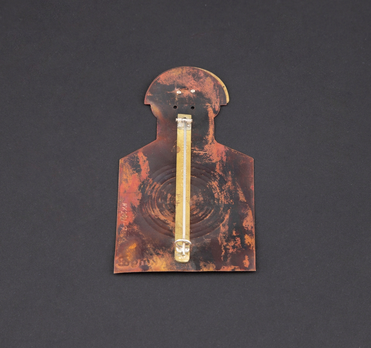 Brosje som forestiller en person med blink på brystet. Personen har gullhat og rødlig kobberkrop. Blink på brystet.