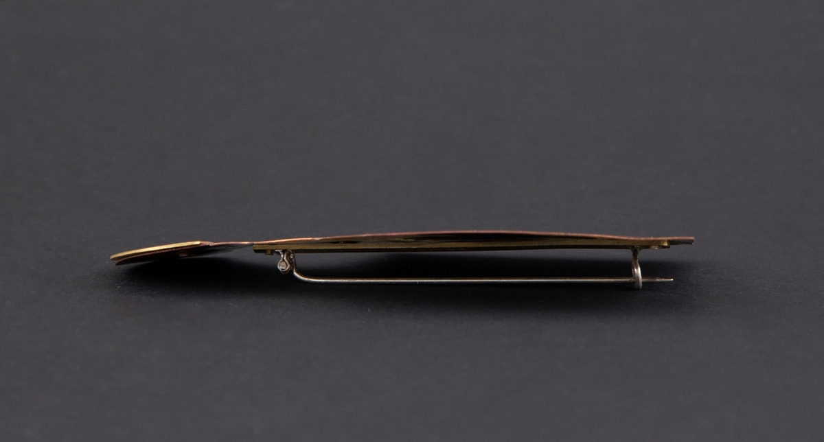 Brosje som forestiller en person med blink på brystet. Personen har gullhat og rødlig kobberkrop. Blink på brystet.