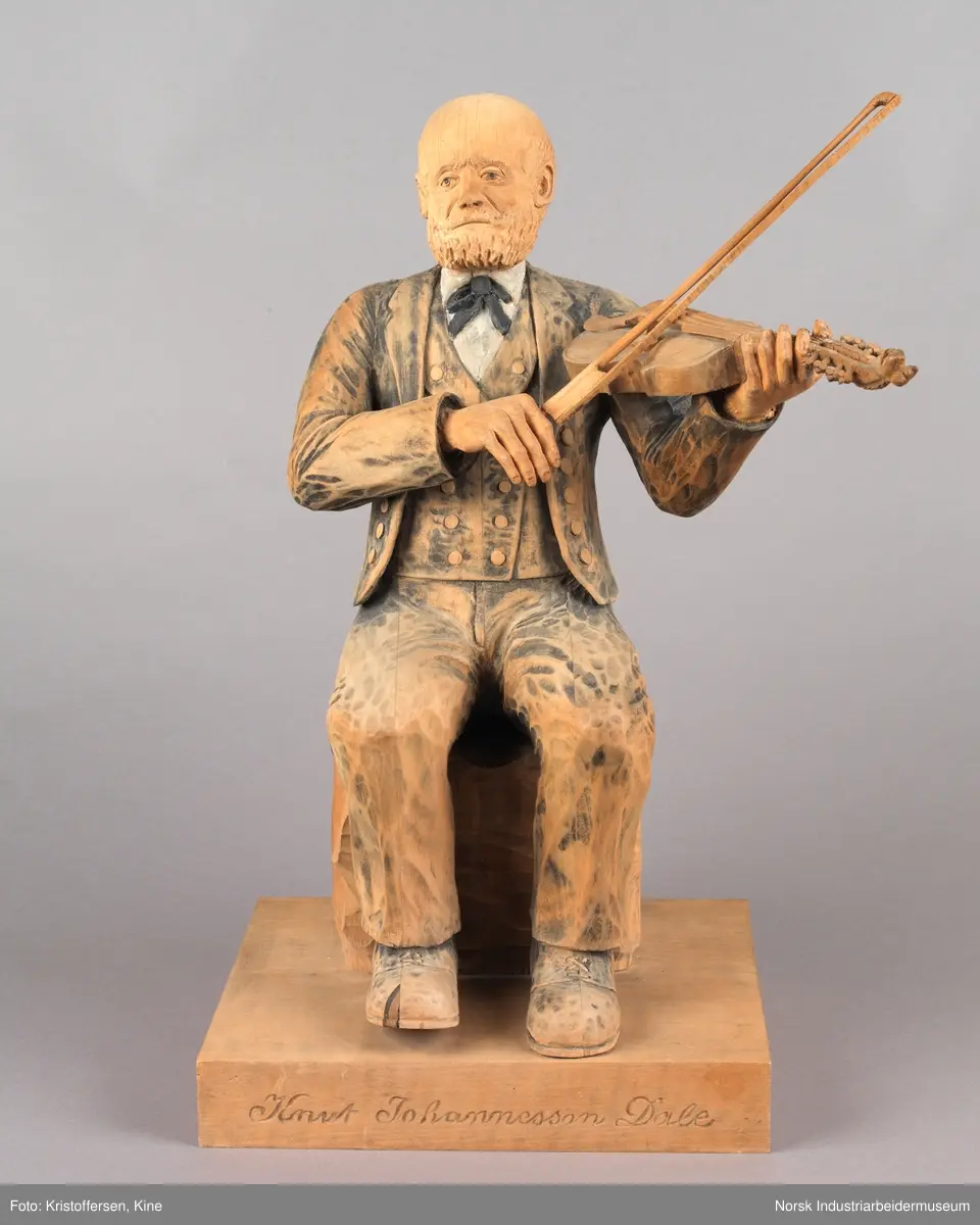 Utskjært treskulptur av spelemannen Knut Johannessen Dahle. Figuren sitter på en kasse med en hardingfele i venstre hånd og en løs bue i den høyre. Han har på seg finklær, sko og sløyfe. Hodet er utskjært med blant annet skjegg, bart og måne. Navnet til Dahle er inngravert på platen.