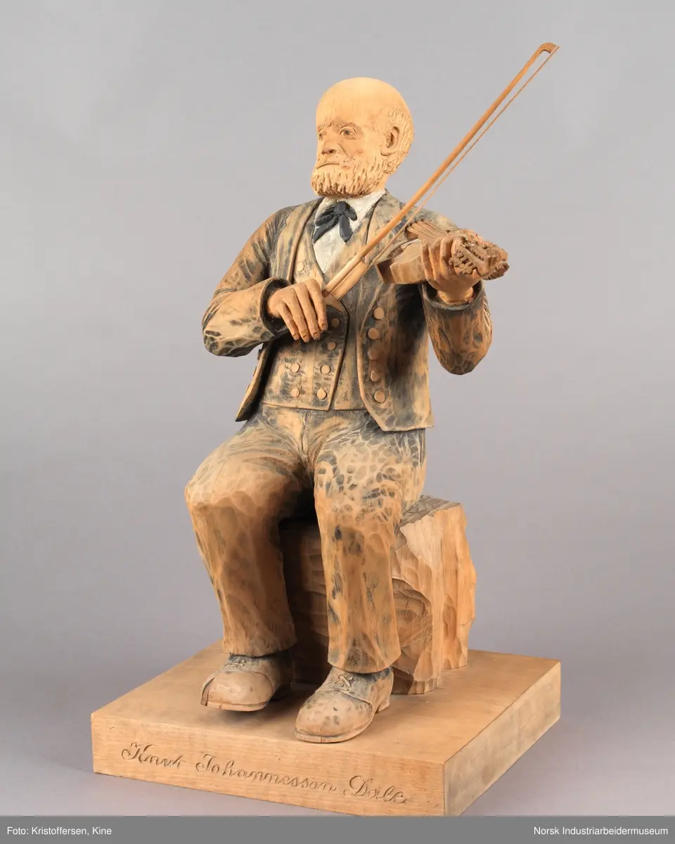 Utskjært treskulptur av spelemannen Knut Johannessen Dahle. Figuren sitter på en kasse med en hardingfele i venstre hånd og en løs bue i den høyre. Han har på seg finklær, sko og sløyfe. Hodet er utskjært med blant annet skjegg, bart og måne. Navnet til Dahle er inngravert på platen.