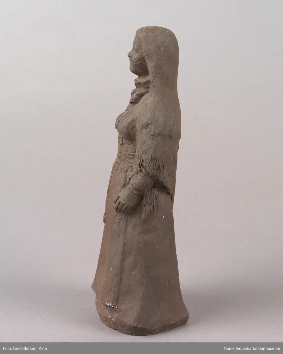 Figur av en kvinne med løst hår iført en bunad. Skulpturen er laget av leire. Bunaden ser ut som en beltestakk fra Telemark. Skulpturen er hul.