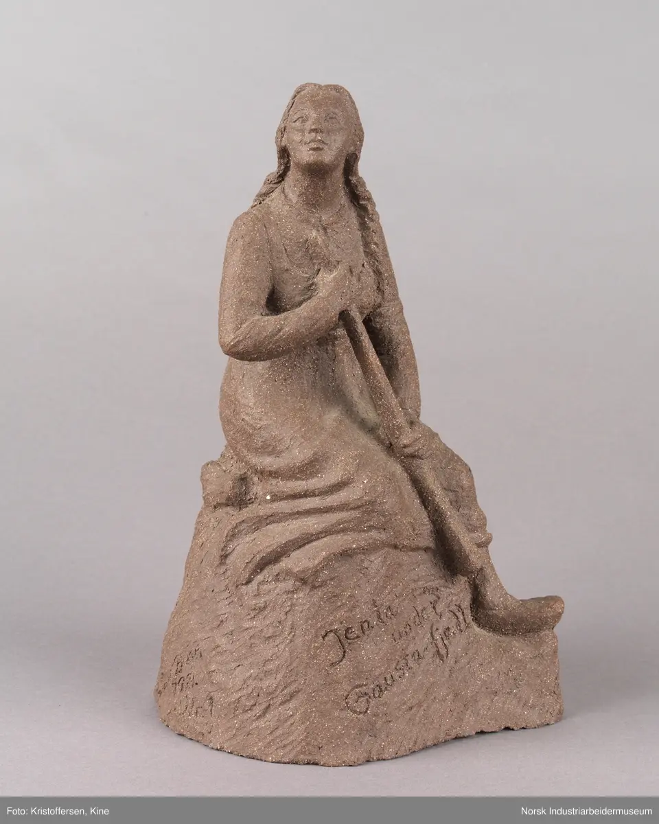 Skulptur laget av leire. Figur, en kvinne med fletter og iført en kjole sitter på en stein med en lur i hendene. Kvinnen vender blikket oppover. Skulpuren er hul.