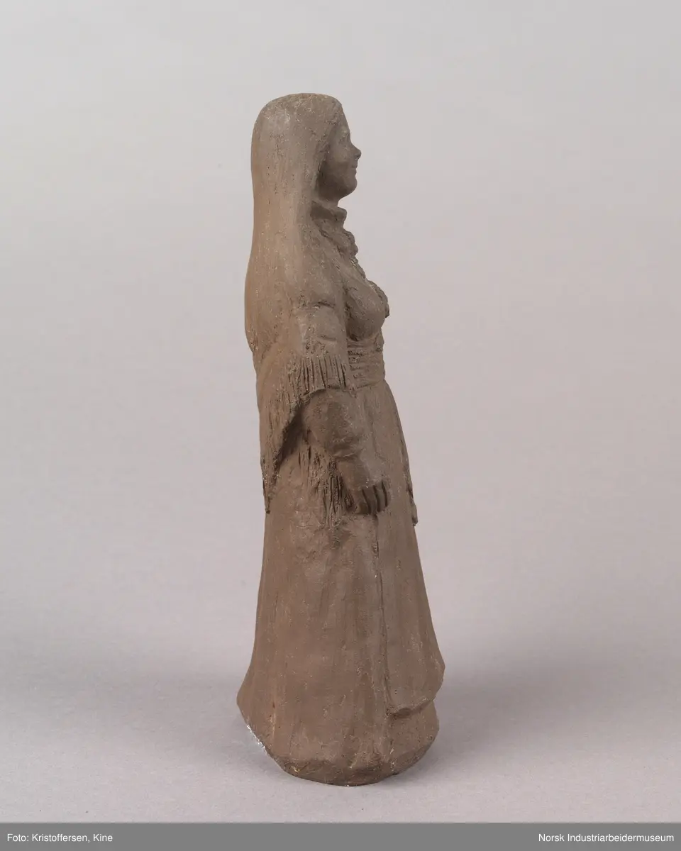 Figur av en kvinne med løst hår iført en bunad. Skulpturen er laget av leire. Bunaden ser ut som en beltestakk fra Telemark. Skulpturen er hul.