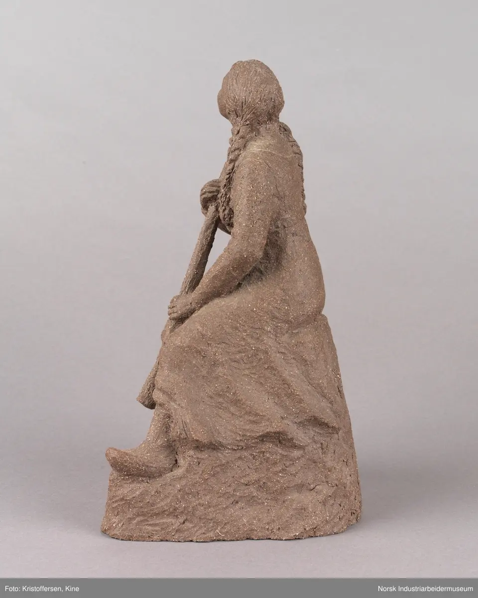 Skulptur laget av leire. Figur, en kvinne med fletter og iført en kjole sitter på en stein med en lur i hendene. Kvinnen vender blikket oppover. Skulpuren er hul.