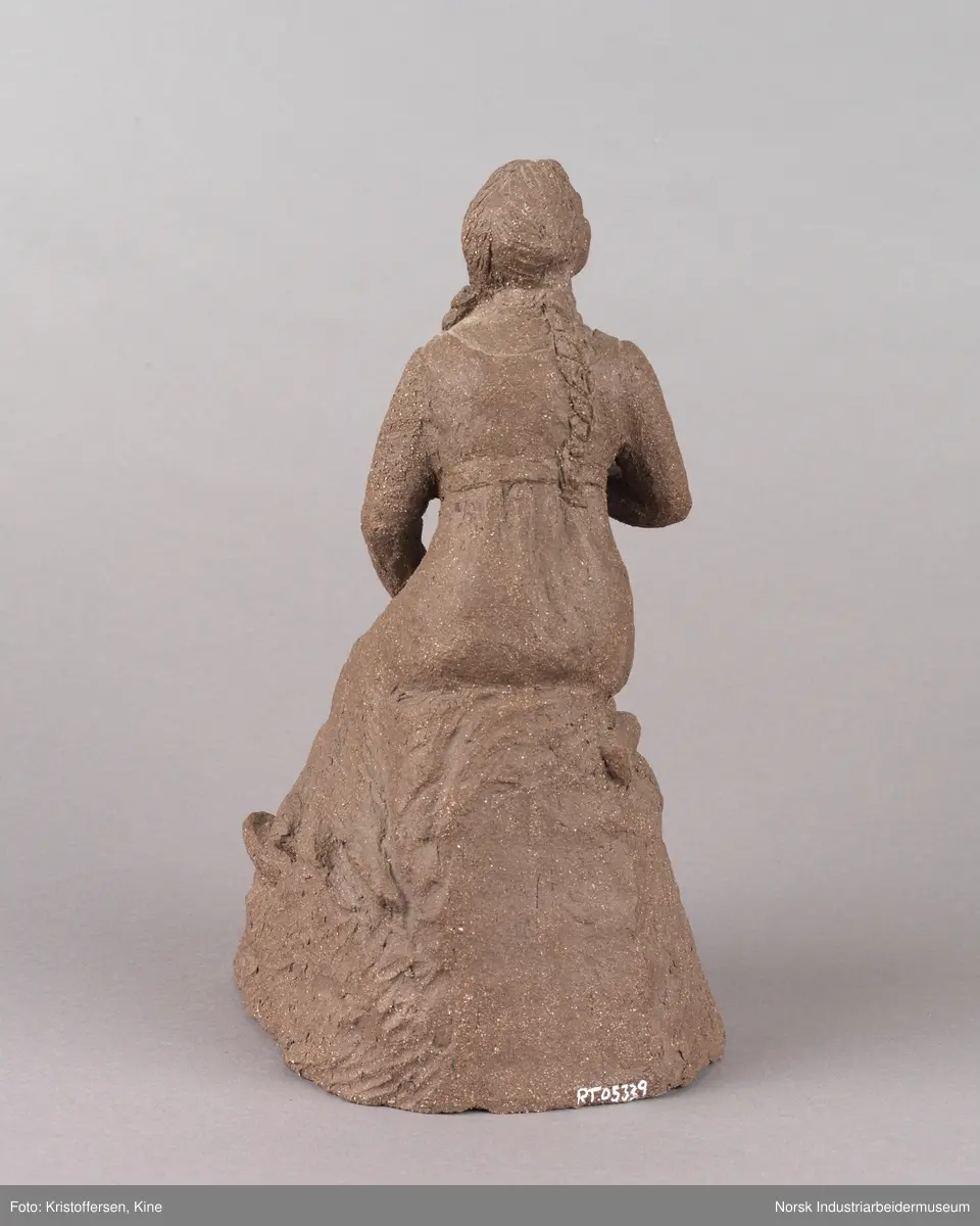 Skulptur laget av leire. Figur, en kvinne med fletter og iført en kjole sitter på en stein med en lur i hendene. Kvinnen vender blikket oppover. Skulpuren er hul.
