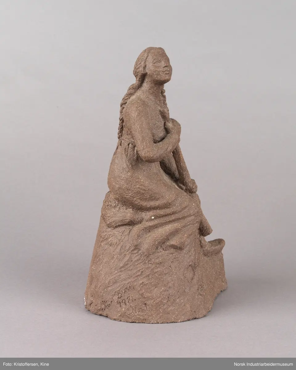 Skulptur laget av leire. Figur, en kvinne med fletter og iført en kjole sitter på en stein med en lur i hendene. Kvinnen vender blikket oppover. Skulpuren er hul.