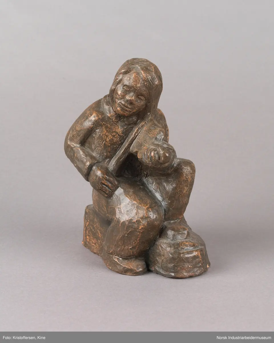 Skulptur, sittende person på sten spiller en fele. Skulpturen er hul i leire og er glassert