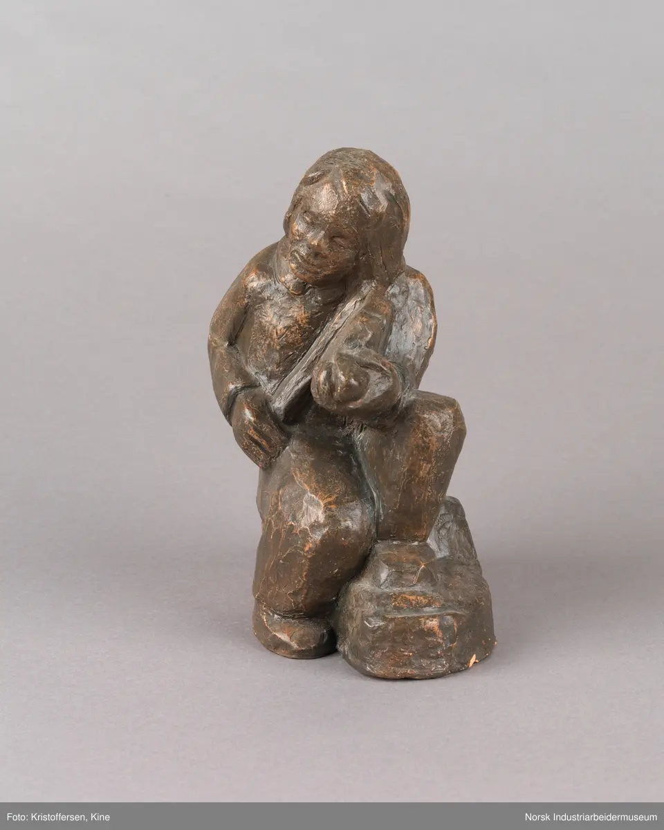 Skulptur, sittende person på sten spiller en fele. Skulpturen er hul i leire og er glassert