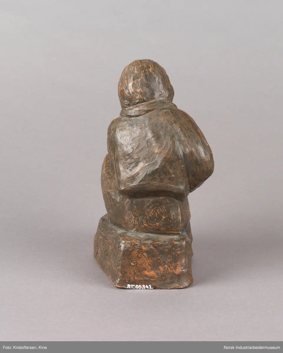 Skulptur, sittende person på sten spiller en fele. Skulpturen er hul i leire og er glassert