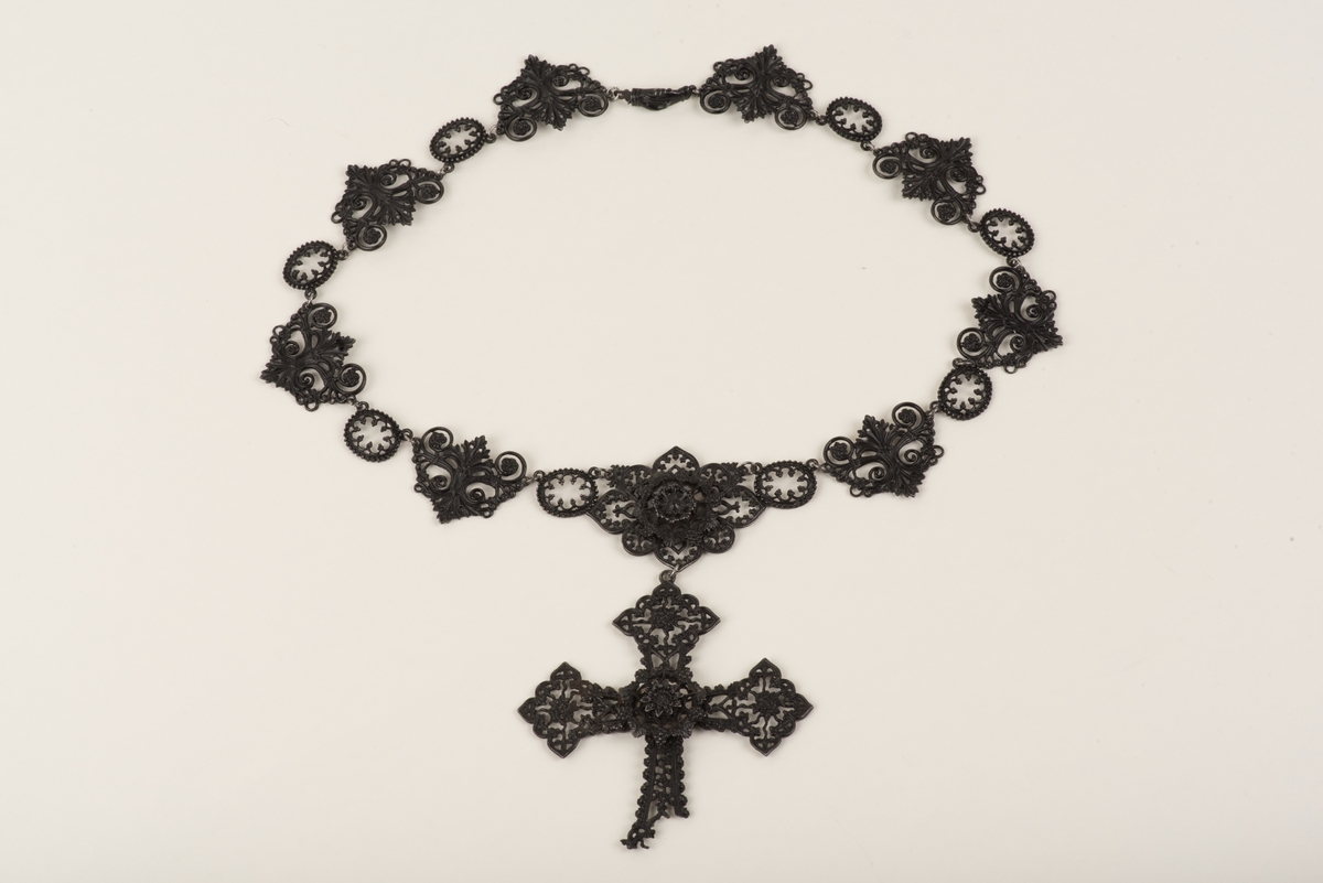 Svart halsband av gjutjärn.
Halsbandet består av omväxlande större och mindre utsirade länkar i nygotisk stil, som är sammanfästa med runda små länkar. Mitt fram hänger ett större likformat kors, som är något skadat. Spännet är utformat som två händer.
