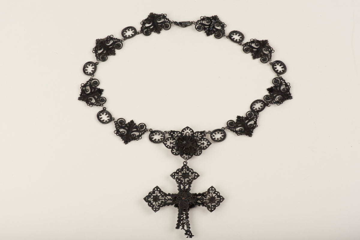 Svart halsband av gjutjärn.
Halsbandet består av omväxlande större och mindre utsirade länkar i nygotisk stil, som är sammanfästa med runda små länkar. Mitt fram hänger ett större likformat kors, som är något skadat. Spännet är utformat som två händer.