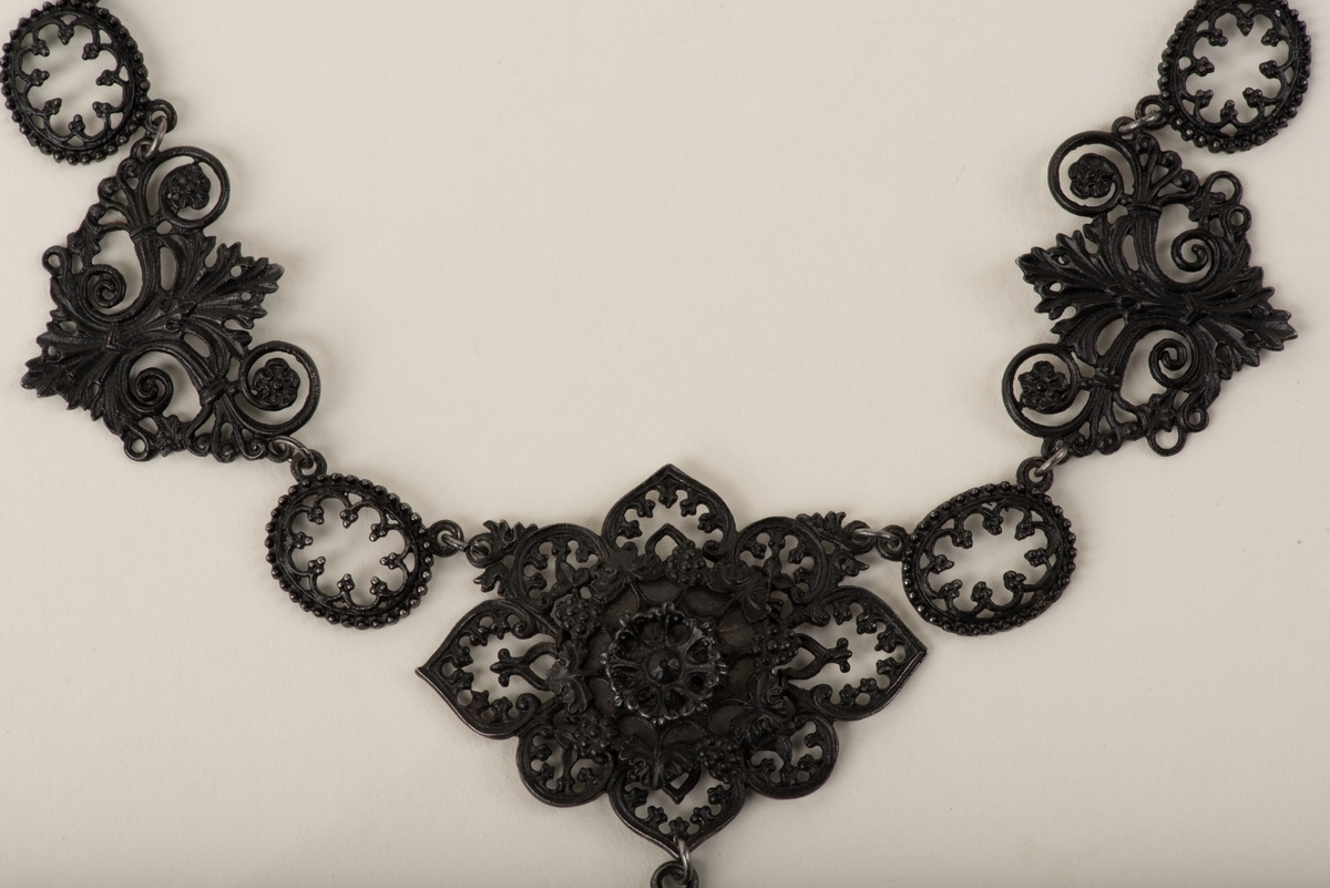 Svart halsband av gjutjärn.
Halsbandet består av omväxlande större och mindre utsirade länkar i nygotisk stil, som är sammanfästa med runda små länkar. Mitt fram hänger ett större likformat kors, som är något skadat. Spännet är utformat som två händer.