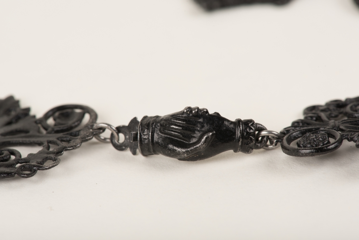 Svart halsband av gjutjärn.
Halsbandet består av omväxlande större och mindre utsirade länkar i nygotisk stil, som är sammanfästa med runda små länkar. Mitt fram hänger ett större likformat kors, som är något skadat. Spännet är utformat som två händer.