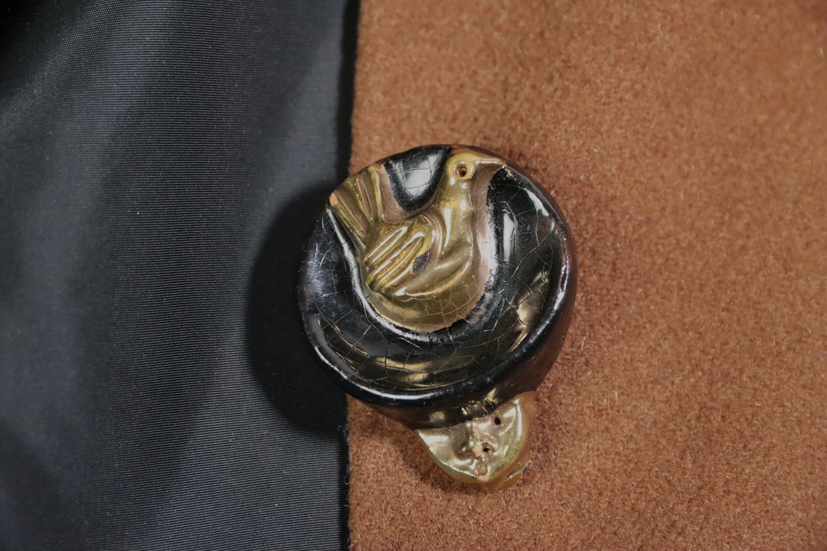 Jacka av ljusbrunt kläde. Figursydd modell med liten svart sammetskrage och accentuerade höfter. Enkelknäppt med stora skulpturala keramikknappar glaserade i mörkt brunt och guld. Knapparna föreställer sittande fåglar. Svart foder med midjeband. Fodret förefaller utbytt. I nacken isydd vävd etikett med loggotype i form av Schiaparellis namnteckning. Etiketten monterad på brett vitt bomullsband, sekundär montering.