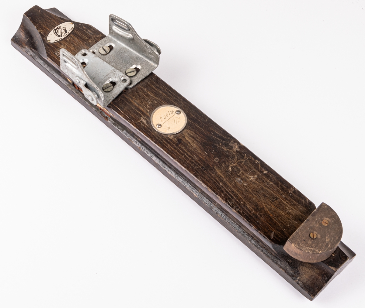 Skidbindningsprov. Vit metallegering på underlag av mörkbrunfärgat trä. På ovasidan Elof Malmbergs firmamärke i metall. Rund metallkantad etikett med skriften "2001 M   N. 7/75" Märke på metalldelen "HOBBY".