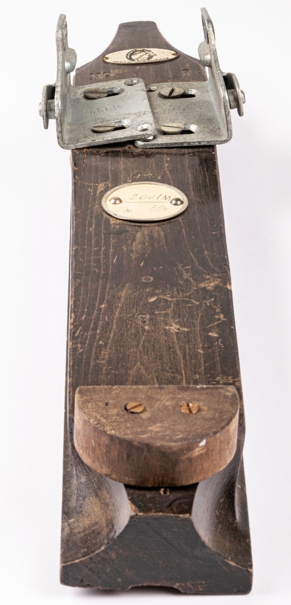 Skidbindningsprov. Vit metallegering på underlag av mörkbrunfärgat trä. På ovasidan Elof Malmbergs firmamärke i metall. Rund metallkantad etikett med skriften "2001 M   N. 7/75" Märke på metalldelen "HOBBY".