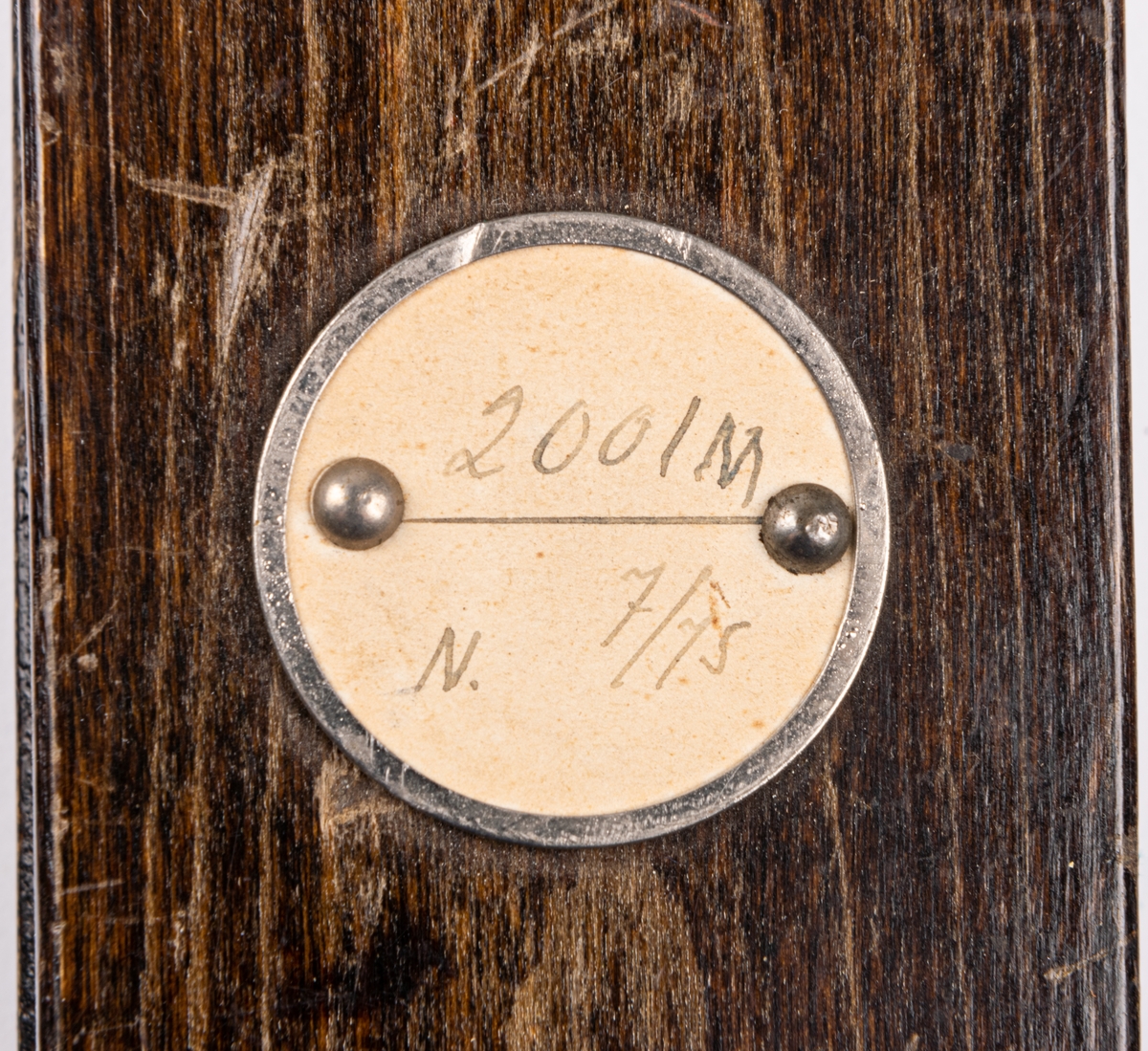 Skidbindningsprov. Vit metallegering på underlag av mörkbrunfärgat trä. På ovasidan Elof Malmbergs firmamärke i metall. Rund metallkantad etikett med skriften "2001 M   N. 7/75" Märke på metalldelen "HOBBY".