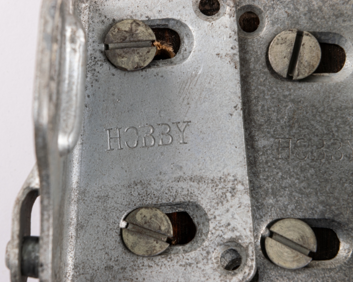 Skidbindningsprov. Vit metallegering på underlag av mörkbrunfärgat trä. På ovasidan Elof Malmbergs firmamärke i metall. Rund metallkantad etikett med skriften "2001 M   N. 7/75" Märke på metalldelen "HOBBY".