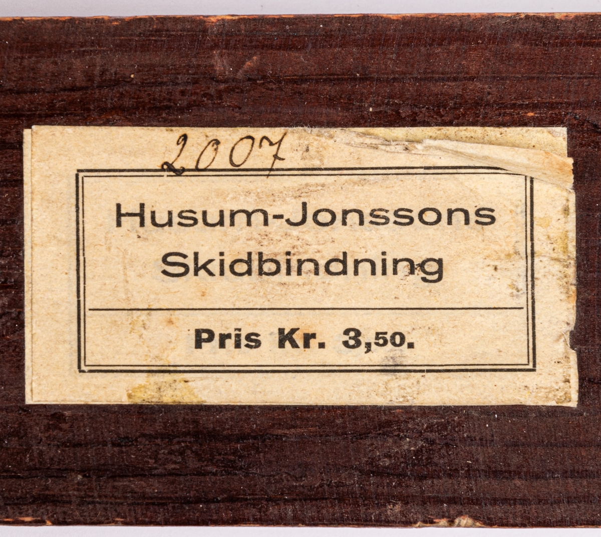 Acc.kat: Skidbindningsprov. Vit metallegering på brunfärgat trä. Läderemmar. Etikett med skrift "2007" och text "HUSUM-JONSSONS SKIDBINDNING PRIS KR. 3,50".