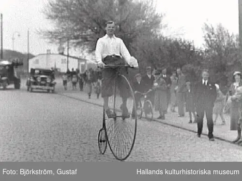 Uppvisning där en man cyklar på en gammal höghjuling, sannolikt på Cykelns Dag i Varberg. Här rullar han förbi åskådrna på Södra vägen vid Rosenfredsskolan. Enligt fotografens liggare är fotot från Sexdagarsloppet det året. Det var en årlig amatörcykeltävling som startades 1924.